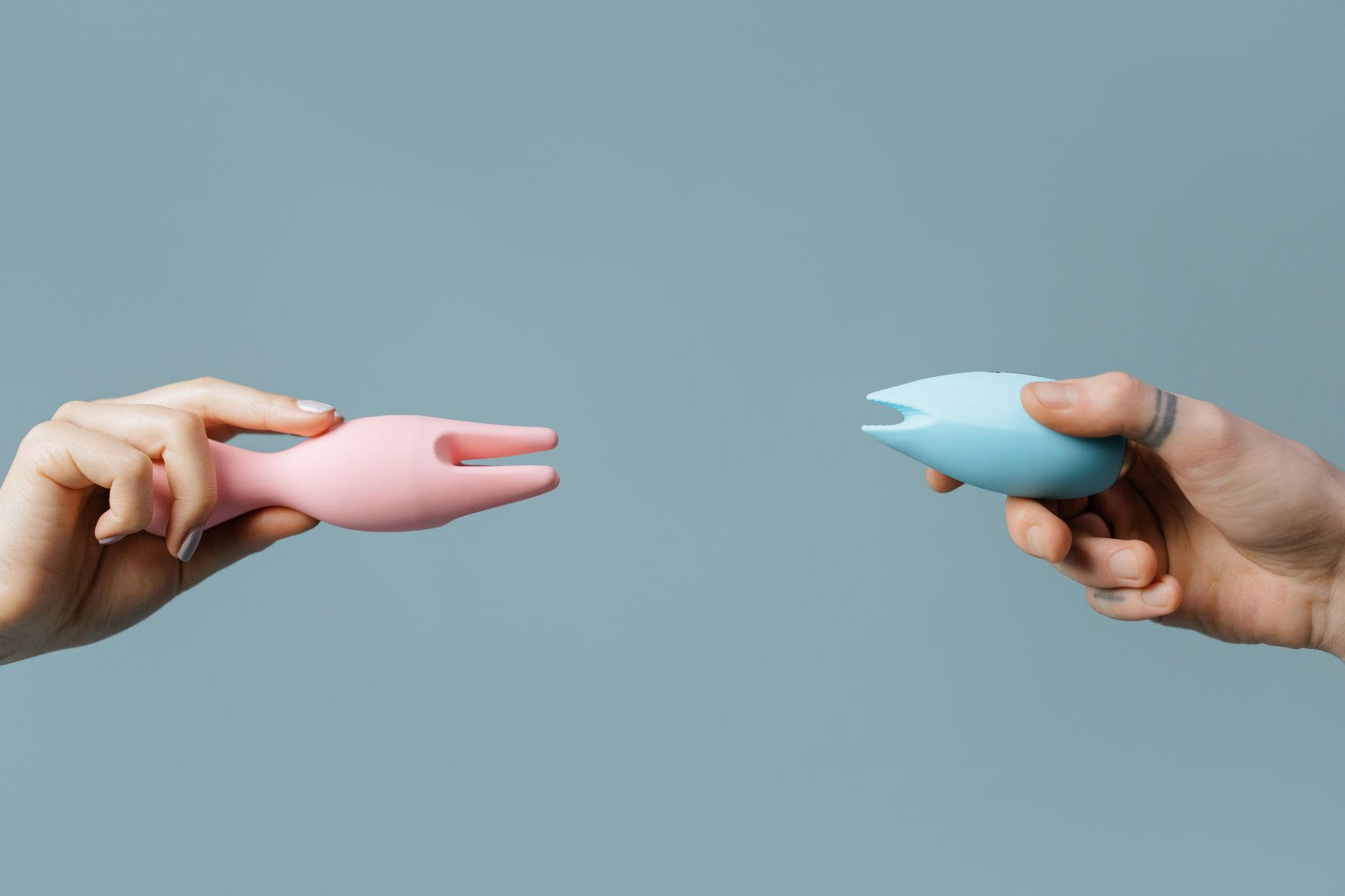 Two hands holding vibrators together against a pastel background, illustrating intimate partnership with pleasure devices.